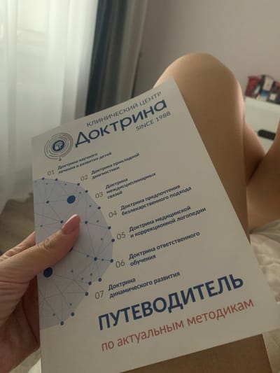 The image shows a hand holding a brochure from a clinic. The brochure has text in Russian, including "КЛИНИЧЕСКИЙ ЦЕНТР ДОКТРИНА SINCE 1988" which translates to "CLINICAL CENTER DOCTRINE SINCE 1988". The brochure lists several points, including "01 Доктрина научного лечения и развития детей" (Doctrine of scientific treatment and development of children), "02 Доктрина прикладной диагностики" (Doctrine of applied diagnostics), and "07 Доктрина динамического развития" (Doctrine of dynamic development). The bottom of the brochure features the text "ПУТЕВОДИТЕЛЬ по актуальным методикам" which translates to "GUIDEBOOK to current methodologies". The background appears to be a bedroom with white curtains and a person's legs visible. There is no information in the image to suggest a specific location in the city of Poritsy, Russia.
