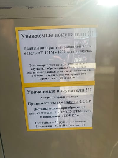 The image is a close-up shot of two informational posters, framed by yellow borders, attached to a textured, light-colored metallic surface, likely a cabinet or locker. The lighting suggests an indoor environment, possibly with overhead lights reflecting on the surface. The posters are printed in black text on white paper and are slightly curled at the edges, indicating they have been displayed for some time.
The top poster, dated 1992, informs customers about a carbonated water dispenser. It states: "Dear Customers! This apparatus is a carbonated water dispenser, model AT-101M - 1992 year of release. This apparatus is one of the few units that has been preserved and is maintained in its original condition, therefore we ask you to handle it with care!!!"
The bottom poster, also in Russian, continues the information for customers regarding the carbonated water dispenser. It reads: "Dear Customers!!! Apparatus for carbonated water. Accepts only USSR coins. Tokens can be purchased at the 'PRODUKTY' store or at the 'BOCHKA' kiosk. 1 kopeck - 25 rubles (carbonated water without syrup). 3 kopecks - 50 rubles (carbonated water with syrup)."
There are no people visible in the image. The posters are the primary subjects, with the metallic surface serving as the background. The context appears to be a commercial or public space where this vintage beverage dispenser is still operational. The mention of "Kablukovo, Russia" is not visually present in the image but is provided as contextual information about the location where this scene might be found.