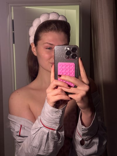 A female subject is depicted taking a mirror selfie indoors in Бакча-Сарай, Russia. The individual, a young adult female, has the right side of her face visible, characterized by a faint smile and a white, textured, bubble-style headband holding back her hair. Her left face is obscured by a smartphone, held in both hands. The phone, which appears silver-toned, is enclosed in a clear, glitter-infused case and features a pink silicone suction-cup array on its reverse. Her fingernails are painted dark red. She is dressed in a light-striped, off-the-shoulder shirt with red trim detailing on the cuffs, exposing her bare shoulders and upper torso. A repeating circular pattern is faintly visible on her upper chest. The immediate background shows a light-colored doorframe to the left and a dark fabric curtain to the right. The scene is dimly lit, with portions in shadow.