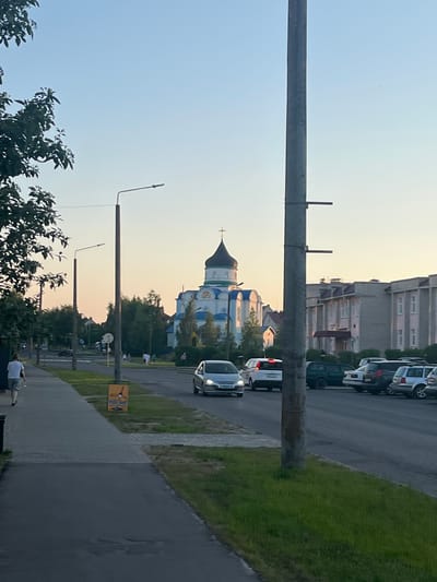 This is a vertical, eye-level, outdoor photograph taken at dusk. The scene depicts a street in Navahrudak, Belarus, with a church and buildings in the background, and a road and sidewalk in the foreground. Several cars are parked along the road, and a white car is driving down it. A few people are visible on the sidewalk, including one sitting on a bench and another standing. Two tall, wooden utility poles with wires extend upwards into the clear sky, which is illuminated by the warm hues of the setting sun. The church, with its distinctive blue and white domes and a cross on top, stands out against the cityscape. The overall atmosphere is peaceful and serene, suggesting the end of a warm day. There is a yellow sign with some text on it in the middle ground, but the text is too small to be legible.