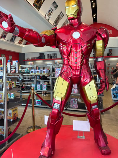An Iron Man statue, approximately 7-8 feet tall, is displayed prominently on a red circular platform in a store. The statue is in its iconic red and gold armor, with its left arm extended forward and its right arm at its side. Behind the statue, shelves are stocked with various merchandise, including toys, action figures, and collectibles. A red velvet rope barrier guides visitors around the statue. In the background, two women are visible, one browsing the shelves and the other carrying a shopping bag. The store appears to be a retail establishment specializing in pop culture and collectible items, possibly a comic book or toy store.