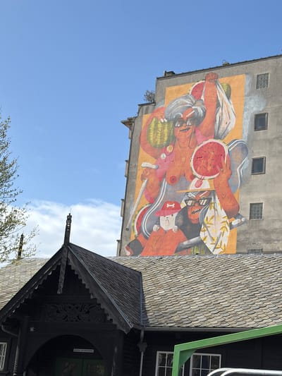 The image shows a mural on the side of a building in Ostend, Belgium, framed by a view of a building and a partly cloudy sky. The mural depicts a vibrant and colorful scene of figures, with watermelon slices and what appear to be surreal or symbolic elements. Below the mural, a building with distinctive dark wooden architectural features is visible, including a roof with layered tiles. A tree is at the bottom of the image. The sky provides a blue backdrop.