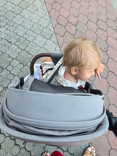 An overhead, eye-level shot captures a baby with blonde hair sitting in a gray stroller. The baby is wearing a beige shirt with a repeating logo pattern and a white t-shirt underneath. The stroller is positioned on a patterned sidewalk made of gray and pink interlocking blocks. The background shows more of the patterned sidewalk and a portion of a dark stroller handle. Two pairs of feet, one wearing white-toed sandals and the other gold sandals with a flower embellishment, are visible at the bottom of the frame, suggesting someone is standing beside or behind the stroller. The lighting appears to be natural daylight.