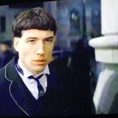 The original image shows a close-up, slightly blurry, head-and-shoulders shot of a young man. He has dark, bowl-cut hair, light skin, and is wearing a white shirt with a dark tie and a dark suit jacket with a thin white trim on the lapel. His gaze is directed slightly to his left. The background is dark and out of focus, with hints of blue and grey, suggesting an indoor or nighttime setting. There is a large, blurred, light-colored object to the right of the man, possibly a column or a structure. The overall lighting is dim. The image does not contain any information or visual cues that would allow for identification of a specific location in Rassvet, Russia.