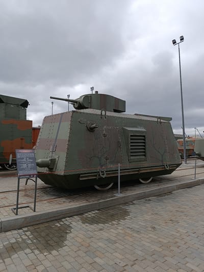 The image depicts the Armored Railcar BN-35, a museum exhibit in the city of Verkhnyaya Pyshma, Russia. The railcar, painted in a camouflage pattern of dark green with lighter green and brown streaks, is positioned on a railway track. It features a prominent turret with a cannon extending forward. Rivets are visible along its hull, and a ventilation grate is present on the side. A sign with text in Russian and English, providing information about the exhibit, stands to the left of the railcar. The surrounding environment includes a paved area with cobblestones, wet in places suggesting recent rain, and other exhibits of military vehicles in the background. The sky is overcast, with visible clouds and a tall lamppost with multiple lights.