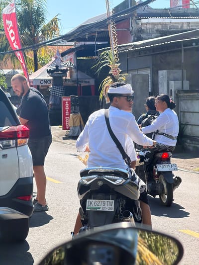 In Kubu, Indonesia, a man with a shaved head wearing a black t-shirt and dark shorts stands beside a white SUV, looking to his left. He is in the foreground to the left of the image. A group of people on motorcycles are in the middle of the road, heading away from the camera. The driver of the first motorcycle, a young man, is wearing a white long-sleeved shirt and a white headscarf. He is looking at the woman riding pillion behind him. The woman is wearing glasses and a white top with a patterned sash around her waist. Her hair is pulled back in a bun. The motorcycle has a license plate that reads "DK 6279 TW 01-29". Another motorcycle is behind them, carrying a passenger with a backpack. The background shows a street with buildings, some with red banners, and palm trees under a bright blue sky. The road is paved and has yellow lane markings. The scene is illuminated by direct sunlight, casting shadows on the road.