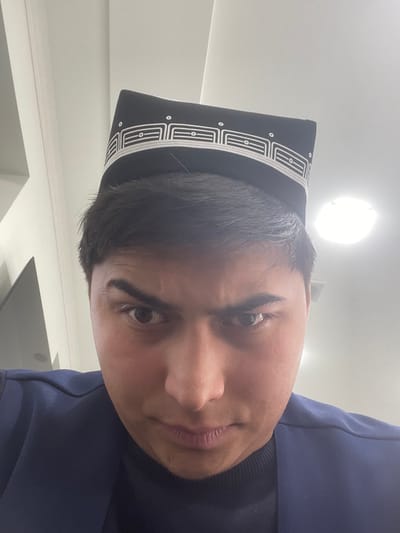 An indoor, close-up image, purportedly from Chinabad, Uzbekistan, displays a male individual from the chest up. He wears a black, square-crowned kalpok with a white geometric embroidered pattern along its lower edge, positioned over dark hair. His facial features include dark eyes with slightly furrowed brows, directing a serious and intense gaze towards the camera. He is clothed in a dark blue jacket or waistcoat over a darker, high-necked undergarment. The background is minimalist, consisting of a plain white wall and ceiling. A circular, flush-mounted light fixture is visible on the ceiling in the upper right, contributing to the bright illumination of the scene. A dark, rectangular object is partially visible on the far left.