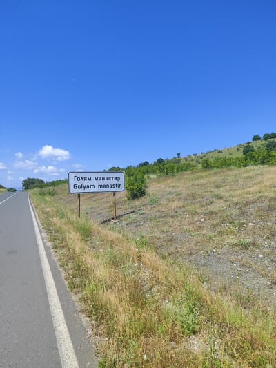 The image shows a sign with Bulgarian and Latin text indicating "Golyam Manastir". The sign is located next to a paved road on a dry, grassy hillside under a clear blue sky. The environment appears to be rural, with sparse vegetation and some trees on the distant hills. The sign is white with black text and is mounted on two wooden posts. The road has a white line marking the edge. The overall scene suggests a location in the countryside of Bulgaria, specifically the village of Golyam Manastir.