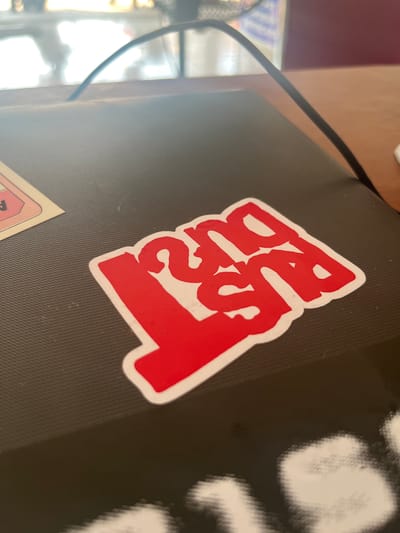 The image shows a close-up of a black, textured surface, likely a laptop lid, adorned with stickers. A prominent red sticker with a white outline features stylized, bold lettering that appears to spell out "BUDDY" or a similar word. The letters are interconnected and designed in a graffiti-like style. To the right of the red sticker, a portion of another sticker is visible, predominantly white with blurry, indistinct shapes that could be text or graphics. The surface itself has a subtle diamond-pattern texture. A black cable is draped across the top left of the image, casting a shadow. In the background, a reddish-brown surface is visible, possibly a desk or table, and a blurred area with bright light suggests a window or a light source in the vicinity. The lighting suggests an indoor setting, likely during the daytime.