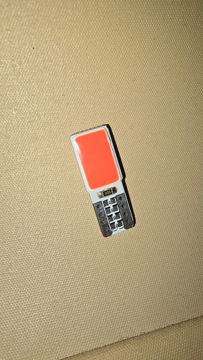 A close-up, overhead shot shows a single, small, orange LED light on a tan, textured surface. The light has a rectangular orange top and a white base with electrical contacts at the bottom. A number "300" is visible on the white base. The tan surface appears to be fabric, possibly upholstery. There are no people or animals present. The image does not contain any information that would indicate its location in Garwolin, Poland.