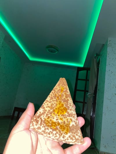A hand is holding a triangular piece of food, possibly a pastry or flatbread, topped with a golden syrup and speckled with brown crumbs. The background features a room illuminated by green LED strip lighting along the ceiling. A ceiling light fixture is visible, along with a textured wall. To the right, a wooden door with glass panes is partially obscured by a light-colored curtain. In the background, a wooden chair or stool is also present. The scene appears to be indoors, likely in a residential setting within the city of Baraki, Algeria.
