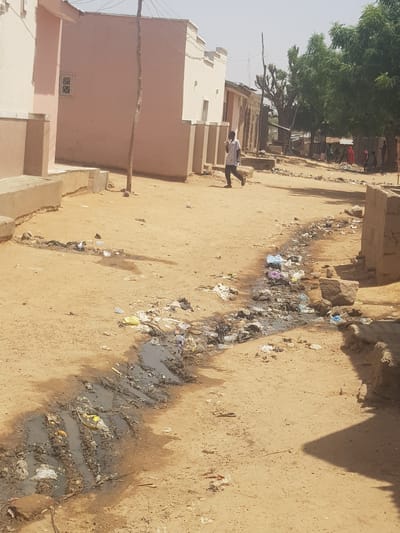 The image shows a dirt street in Dutsinma B, Nigeria. Litter and debris are scattered along a channel running down the center of the street. Water accumulates in the channel, creating a muddy, unsanitary appearance. Buildings line the side of the street, with visible walls and roofs constructed from clay and concrete. A person walks along the street toward the background. A tree line is visible further in the background. The environment appears dry and dusty.