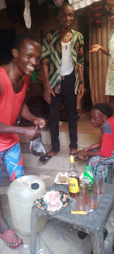 This media file depicts a casual social gathering in Ohafia, Nigeria, likely in an indoor or covered outdoor area such as a porch or veranda. The scene features four individuals and various objects on a low table.
In the left foreground, a young, dark-skinned man with a broad smile wears a red tank top and blue patterned shorts. He is actively engaged, holding a clear plastic bag containing a dark liquid, possibly preparing to serve a drink. His right foot, clad in a red sandal, is visible. Standing prominently in the center background is another young, dark-skinned man, dressed in a white undershirt, black trousers, and a distinctive green, yellow, and black patterned jacket, typical of an ankara print. He wears a dark pendant necklace and black flip-flops, with a neutral expression.
To the right foreground, an older, dark-skinned woman is seated on a low stool. She wears a red top with a dark panel, a patterned skirt or wrapper, and a dark headwrap. Her attention appears to be directed downwards, towards her lap or the ground in front of her. Partially visible in the far right background, behind a patterned curtain, is another person's raised hand.
The central focus of the foreground is a dark, low, rectangular table laden with items. These include a bottle prominently labeled "501" (likely a spirit), a green plastic bottle (possibly soda or water), and several transparent glasses. A small, floral-patterned tray on the table holds snacks or food items, alongside stacks of Nigerian Naira banknotes, suggesting a possible transaction or gift. A yellow bottle opener is also present. Adjacent to the table is a large, translucent white plastic jerrycan with a black cap.
The overall atmosphere suggests an informal social occasion. The man on the left appears happy or amused. The attire, the "501" brand bottle, and the Nigerian Naira notes are consistent with the specified Ohafia, Nigeria, location context. The lighting indicates it is likely daytime, but the area appears to be shaded.