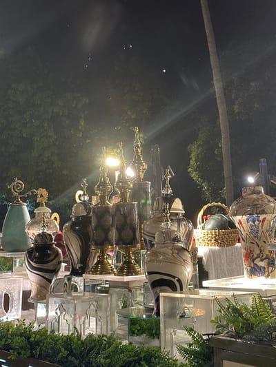 The image displays a brightly lit exhibition or retail display of decorative vases and home decor items against a dark, nighttime outdoor background. The photograph is oriented 90 degrees counter-clockwise from a standard upright view.
In the foreground, a multi-tiered display showcases numerous ornate vases and jars. The items are arranged on clear, possibly acrylic or glass, shelving units, some of which feature decorative cut-out patterns. To the far right, lush green foliage, resembling ferns, adds a touch of natural accent to the display.
The decorative pieces themselves vary greatly in style and material. Towards the upper right, a large, intricately patterned ceramic vase features a blue and orange floral design. Below this, and continuing across the display, are rows of distinct vases. Many are characterized by glossy finishes, with patterns such as black and white stripes, mottled marble-like effects, and geometric designs on darker, cylindrical bodies. The majority of these vases incorporate metallic, gold-colored bases, necks, and decorative finials. Several of the finials are adorned with clear, crystal-like elements, adding to their luxurious appearance. One unique piece towards the bottom left of the display is a teal or greenish vase with an elaborate golden, swirling handle or embellishment. A woven basket is also subtly visible behind some of the vases in the upper right section.
The display is illuminated by multiple warm, yellow artificial lights, some of which are visible as bright glows or cause significant light streaks and reflections, particularly on the shiny surfaces of the vases and metallic elements. These lights create a vibrant and inviting atmosphere for the exhibited items.
The background is predominantly dark, indicating that the scene takes place at night. Dark silhouettes of trees are visible, with one prominent, slender tree trunk (likely a palm) extending diagonally across the upper left portion of the image. Scattered within the dark background are several small, faint white lights, which could be distant light sources or atmospheric phenomena.
The overall impression is one of an elegant nighttime display, possibly at a market, outdoor shop, or special event, showcasing ornate decorative objects. No visible text is present in the image.
