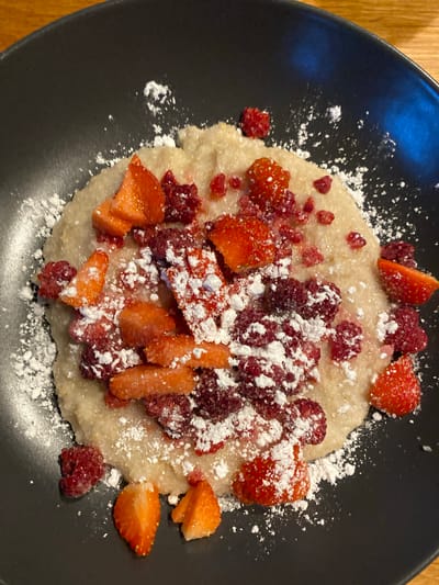 A plate of porridge topped with chopped strawberries and raspberries, sprinkled with powdered sugar, is presented on a dark gray plate. The plate is placed on a wooden surface. The location in Lommedalen, Norway, is not discernible from the image.