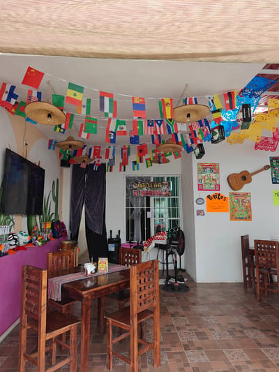 This image depicts the outdoor seating area of a restaurant or cantina, likely in Cancún, Mexico, judging by the festive decorations and cultural motifs. The space is adorned with strings of international flags, sombreros serving as light fixtures, and colorful papel picado banners. A large flat-screen television is mounted on a wall decorated with a cactus mural. Several wooden tables and chairs are arranged on a tiled floor. On one table, a cloth runner with a striped pattern is laid out, holding napkins and what appears to be a small menu or sign. In the background, a guitar hangs on the wall, accompanied by several framed posters, some featuring what looks like soccer imagery, hinting at a sports bar or themed establishment. The overall atmosphere is lively and casual, suggesting a place designed for socializing and enjoying food and drinks.