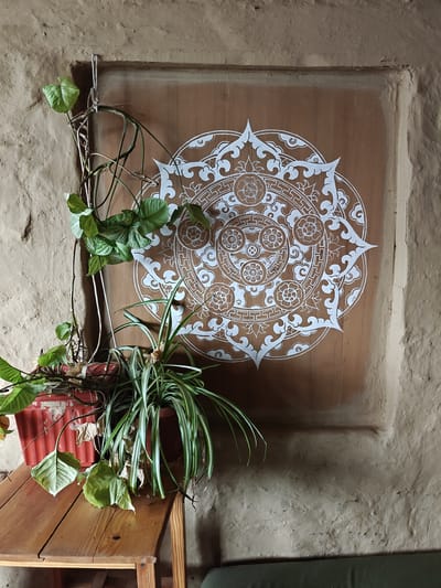 In Prini, India, a close-up shot captures a detail of a wall adorned with a white mandala pattern on a brown wooden panel. To the left, a red planter holds two types of plants: a vine with heart-shaped leaves cascading down and a spider plant with long, striped leaves. The wall itself is textured, appearing to be made of rough plaster or adobe. A wooden table sits in the foreground, supporting the planter. The overall impression is one of rustic, natural decor.