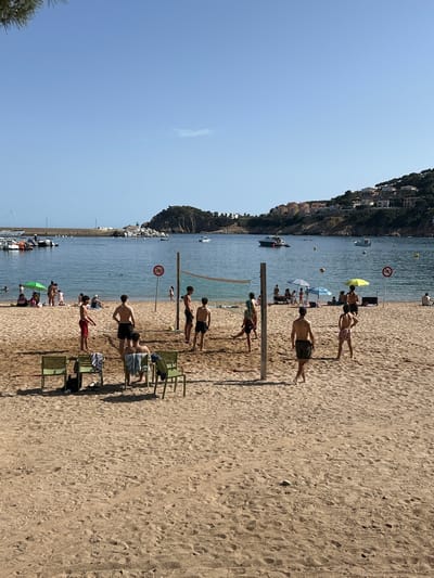 In the image, a beach volleyball game is in progress on a sandy beach. Several young men are actively playing, with one in mid-air appearing to spike the ball. Spectators are visible in the background, some sitting and some standing, with beach umbrellas providing shade. Beyond the beach, the calm blue sea stretches out, dotted with a few boats. In the distance, a hilly coastline is lined with houses, suggesting a town or village. The sky is clear and blue, indicative of a sunny day. The overall scene depicts a lively and relaxed atmosphere at a coastal recreational area.