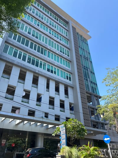 A low-angle, eye-level outdoor medium shot captures the facade of a modern, multi-story building with an open-air parking garage on its lower levels. The building, located in Makassar, Indonesia, features a combination of grey concrete and glass panels. The upper floors have rows of evenly spaced, rectangular windows with light teal-tinted glass. The parking garage levels reveal open rectangular cutouts with vegetation growing in the spaces. A grey, diagonal patterned panel is visible on one section of the building's corner. A large signpost with directional arrows and text in Indonesian is positioned in front of the building, along with lush green foliage and palm trees. A black car is parked in front of the building's entrance, which is partially obscured by the foliage. The sky above is clear and bright blue, with a tree on the right side of the frame casting shadows on the building. A white circular object, possibly a satellite dish, is mounted on one of the parking garage levels. A red and white circular no-smoking sign is affixed to the wall near the building's entrance.