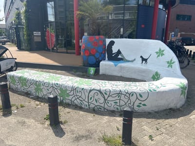 An outdoor shot captures a curvilinear white concrete bench decorated with green ivy-like vines and leaves. A silhouette of a person reading is painted on the bench, accompanied by a small black cat silhouette. Adjacent to the bench is a large, cylindrical planter with a red and blue polka-dot pattern, containing a palm tree. In the background, a modern building with large glass windows is visible, featuring an entrance with a large graphic of a red raspberry on the glass door. A bicycle rack filled with bicycles is situated to the right of the building. The scene is set on a paved area with some scattered weeds and is illuminated by sunlight, casting shadows from the bollards lining the edge of the bench. This location is identified as the Bio Science Park in Leiden, Netherlands.