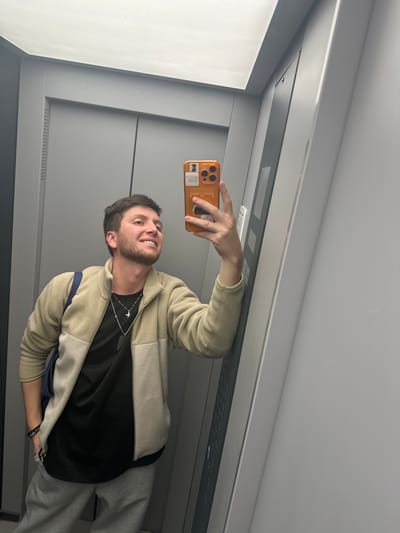 A young man with short brown hair and a beard takes a selfie in an elevator. He is wearing a beige and cream zip-up fleece jacket over a black t-shirt. He has a silver necklace with a cross pendant and a black beaded bracelet on his left wrist. He is also carrying a dark blue bag over his left shoulder. The elevator walls are grey and appear to be metal. The man is smiling and looking at his phone, which has an orange case. The image is taken from a slightly low angle. There are no other people visible in the image. The man is standing and appears to be posing for the photo. There is no indication of the specific location within the city of Мирный, Russia, other than it is an elevator.