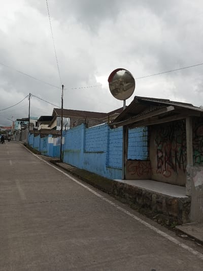 This image depicts a street scene in Melakudikadu, India, under an overcast sky. The concrete road, marked by a white line on its right edge, ascends slightly and curves gently to the left in the distance. To the right of the road, a long brick wall, painted light blue and topped with barbed wire in sections, runs parallel to the street. Adjacent to the road and against the blue wall, a small, open-front shelter with a corrugated metal roof and a stone-clad base is present; its interior back wall is covered in red and green graffiti. Above and behind this shelter, a large, circular convex traffic mirror is mounted on a pole, reflecting a blurred image of distant structures. Utility poles with multiple overhead wires are visible along the street. In the far distance on the left, a motorcycle with at least one rider can be seen heading away from the viewer. Buildings with various roof types are partially visible behind the blue wall and further down the street.