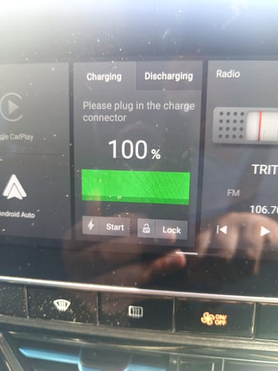 The image shows a car's infotainment screen displaying charging status for an electric vehicle. The screen indicates "Charging" is selected, and the battery is at "100%". Below the charging percentage, a green bar visually represents the full charge. To the right of the charging status, "Discharging" and "Radio" are also visible as selectable options. Further to the right, a radio tuner displays "TRIT FM" and the frequency "106.70". At the bottom of the screen, there are icons for "Start" and "Lock", along with navigation arrows. The car's dashboard controls are visible at the bottom of the image, including a windshield wiper icon and a hazard light button illuminated in orange with "ON/OFF" text. The screen is dusty. The image was taken in Pyrgos Community, Cyprus, likely inside a vehicle.