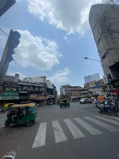 The image captures a street intersection in Ahmedabad, India, under a partly cloudy sky. A traffic light is visible on the left, casting a shadow. In the foreground, a green and yellow auto-rickshaw is stopped at a pedestrian crossing. The street is paved with asphalt, and white stripes demarcate the pedestrian crossing. Various shops and buildings line the sides of the street, displaying signage for businesses like "OPPO" and "DIMPLE MOBILE." Several other vehicles, including cars and motorcycles, are present on the road, with some people walking on the sidewalks. A tall, somewhat dilapidated concrete building with visible windows stands on the right side of the intersection, and a street lamp is mounted on a pole nearby. Billboards with advertisements are also visible on some of the buildings. The scene depicts a typical urban street scene with moderate traffic and commercial activity.