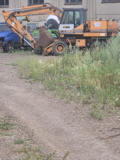 An orange Case Poclain 888B excavator is parked on a gravel lot in front of a building. The excavator has a blue truck parked to its left, and is surrounded by tall green grass and weeds. The building appears to be industrial or commercial. The scene is outdoors during daylight. The image shows the excavator from a side angle, with the front loader arm extended. The excavator has the model number "888B" printed on its side. There are no people visible in the image. The location is identified as Novoişimsk, Kazakhstan.