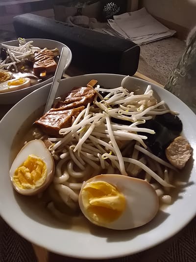 A close-up shot shows two bowls of ramen on a surface with what seems to be cushions or pillows to the side. The ramen bowls contain noodles, halved boiled eggs with cooked yolks, bean sprouts, dark-colored mushrooms, and pieces of what appears to be braised pork belly or similar meat. A pair of metal tongs rest on one of the smaller bowls. The lighting suggests an indoor setting, possibly a home environment, but there's no direct evidence of the location being in Aubervilliers, France.