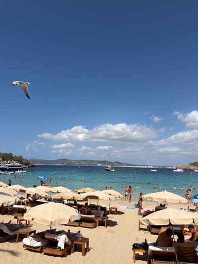 The image depicts a bustling beach scene in Sant Josep de sa Talaia, Spain, under a clear blue sky with scattered white clouds, suggesting a bright, sunny day. The time appears to be daytime, likely afternoon, given the strong shadows cast by the beach umbrellas and furniture.

The foreground and middle ground are dominated by a sandy beach populated with numerous beachgoers. Several beach umbrellas, some light beige and others darker, are set up, providing shade. Beneath these umbrellas are lounge chairs and small tables, occupied by people relaxing and socializing. The beach is lined with sunbeds and wooden furniture, indicating a commercial beach establishment.

In the turquoise water, many people are swimming and enjoying the sea. A few boats, including a white sailboat and a larger boat with a blue canopy, are visible near the shore and further out at sea. The coastline in the background features hills with some vegetation and buildings, characteristic of a Mediterranean setting.

The image captures a lively atmosphere of a vacation destination, with people engaged in leisure activities. The overall mood is relaxed and festive, typical of a summer beach day. A single seagull is captured in mid-flight against the vast blue sky in the lower left portion of the image. There is no visible text in the image.