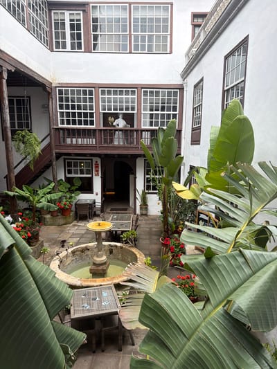 This image depicts a charming inner courtyard in San Cristóbal De La Laguna, Spain. The scene is characterized by traditional architecture with white stucco walls and dark wooden window frames, featuring numerous multi-paned windows. Lush, large-leafed plants, likely banana plants, dominate the foreground and midground, their vibrant green leaves creating a tropical feel against the old-world structures.
In the center of the courtyard, a weathered stone fountain with a yellow basin is a prominent feature. Surrounding the fountain are potted plants with red flowers and various greenery, adding color and life to the space. Several outdoor seating areas with wicker furniture and glass-topped tables are visible, suggesting a relaxed atmosphere, perhaps a cafe or a hotel courtyard.
A second-story balcony with wooden railings and a statue of a person holding flowers is visible on the left side of the building. The lighting suggests it is daytime, with soft, diffused light, possibly indicating an overcast sky or shaded conditions within the courtyard. The overall impression is one of tranquility and historical charm, with a touch of exotic flora.
One visible sign, with red and white lettering, is located near a doorway, but the text is not clearly legible. The scene conveys a sense of calm and provides a glimpse into the architectural style and natural beauty of the location.