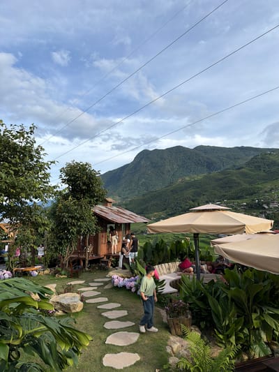 The image depicts an outdoor cafe or resting area set against a backdrop of lush green mountains. Several people are visible, some seated and others standing or lying down. In the foreground, a young man in a light green t-shirt and dark jeans is lying on the grass. Further back, a group of people are seated on a wooden bench, with a child lying down nearby. There are large, light-colored umbrellas providing shade. The setting includes a small wooden structure, possibly a cafe or shop, with a corrugated metal roof. A pathway made of large, flat stones leads through the grassy area. Various plants, including large leafy varieties and flowering bushes, are scattered around. In the distance, the mountains are covered in dense vegetation, and some houses or structures can be seen on the slopes. Overhead, power lines stretch across the sky, which is partly cloudy with patches of blue. The scene appears to be during the daytime, with natural light illuminating the area. The overall atmosphere seems relaxed and leisurely, suggesting a tourist spot or a peaceful retreat. The location is identified as Tả Van, Vietnam, which aligns with the mountainous and terraced landscape often associated with that region. No visible text is present in the image.