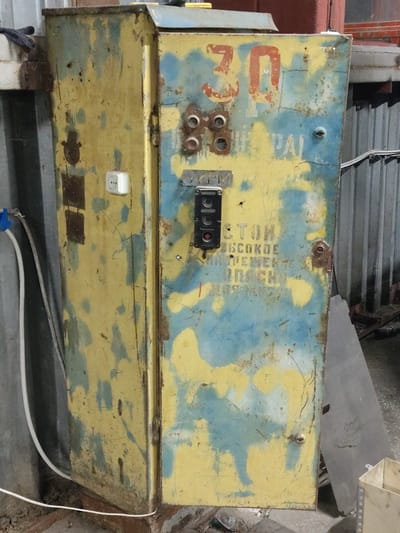 The image shows a heavily weathered, industrial-style metal cabinet painted yellow and blue. The paint is chipped and peeling, revealing rust underneath. A red number "30" is painted on the upper portion of the cabinet. There is a black electrical control panel mounted on the front, with a red emergency stop button visible. Below this, text is stenciled in yellow, though much of it is obscured or illegible due to the damage and paint. Some of the visible text appears to be in Cyrillic, possibly indicating a warning or operational instruction. A white electrical outlet is mounted on the left side of the cabinet. The cabinet is positioned outdoors or in a semi-enclosed industrial space, with corrugated metal siding visible in the background. Electrical conduits and cables are also present. The ground is covered in debris, including dirt and possibly construction materials. The overall impression is one of disuse and decay in an industrial setting. The location is identified as Dangir, Uzbekistan.