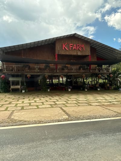 The image shows the exterior of a two-story wooden building with a large, sloped roof. Red lettering on the upper facade spells out "K' FARM". A dark awning covers the lower level, which features large windows and a balcony on the second floor. Red lanterns are suspended from the building, and potted plants are visible on the ground level. The building is set on a paved area made of large, square concrete blocks with grass growing between them. A road with a white line runs along the bottom of the image. The scene is likely in Tu Póh, Vietnam, based on the architecture and signage.