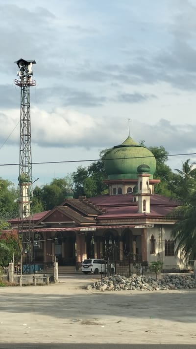 The image shows a mosque in Bulo Gogo, Indonesia. A tall metal tower with loudspeakers is located to the left of the mosque. The mosque has a large green dome and a smaller green dome on a minaret. The building features a red tiled roof and arches. A white car is parked in front of the mosque. The foreground is a dirt or gravel area. Trees and palm trees are visible around the mosque. The sky is cloudy.