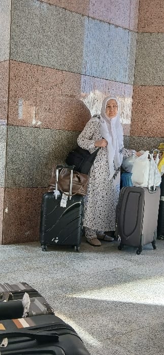 A female subject stands upright in what appears to be a public or semi-public area in Medina, Saudi Arabia, characterized by a wall composed of large, rectangular, variegated tiles in shades of gray, light brown, and pale blue. She is wearing a light-colored, long-sleeved dress with a dark speckled pattern and a white hijab. A black shoulder bag is slung over her left arm. She is positioned amidst multiple pieces of luggage: a black hardshell suitcase with a brown leather tote bag on top is to her left, and a gray hardshell suitcase topped with a white plastic bag is to her right. Another black suitcase is partially visible in the lower foreground. The floor is light-colored and speckled, possibly concrete or terrazzo. Strong, directional lighting from the right casts shadows of the subject and her luggage onto the wall and floor. The subject's gaze is directed slightly to her right, towards the viewer's left.