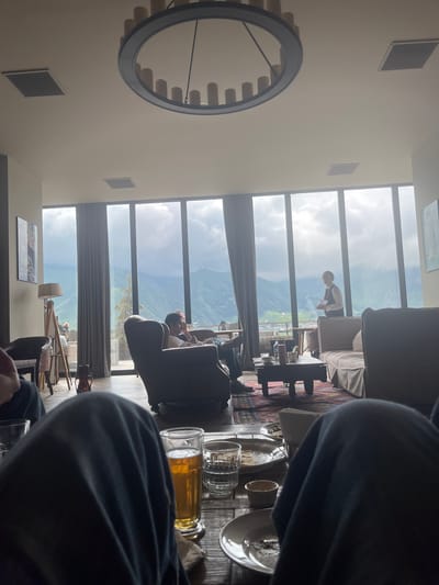 This image captures a dimly lit, cozy interior of a restaurant or lounge in Stepantsminda, Georgia, with a breathtaking view of the surrounding mountains visible through a large window. In the foreground, the dark fabric of clothing obstructs the view of two people. The middle ground reveals a seating area with plush armchairs, a sofa, and a coffee table laden with drinks and plates. One person is seated in an armchair, appearing to be in conversation with another individual who is partially visible. A server is standing near the window, holding a document. The background is dominated by a panoramic vista of lush green mountains under a dramatic, cloudy sky, suggesting a serene, natural setting. A distinctive, large chandelier hangs from the ceiling.
