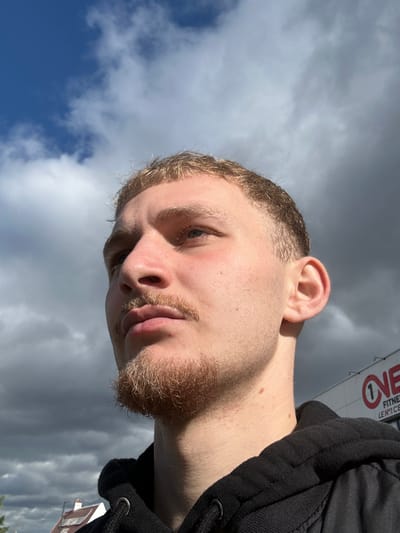 The image depicts a young Caucasian male outdoors under a partly cloudy sky in Fegersheim, France. The subject, seen from the upper torso, has short blonde hair with faded sides, a blonde mustache, and a matching goatee. His head is angled slightly to the right, with his gaze directed upwards and to the viewer's left. Strong direct sunlight illuminates the left side of his face, casting a shadow under his chin. He wears a black hooded sweatshirt with visible drawstrings. In the background, to the subject's right, a commercial building displays a red and white 'ONE' logo with a '1' inside the 'O', followed by the partially legible text 'FITNESS' and 'LENT 'C'', indicating 'ONE FITNESS CENTER'. To the lower left, a portion of a residential building's roof and white wall are visible.