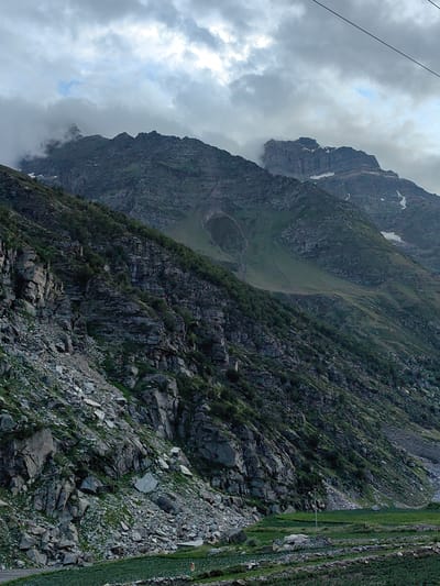 This is a vertical, medium shot photograph of a mountainous landscape with a cloudy sky. The foreground features a terraced agricultural field with small green plants and stone walls. The midground is dominated by a steep, rocky mountainside covered in sparse vegetation, including small trees and shrubs. The rocks are varied in size and texture, with some large boulders and scree slopes. In the background, several large, rugged mountain peaks rise, some of which are partially obscured by clouds and mist. Patches of snow are visible on some of the higher peaks. The sky is overcast with heavy, grey clouds, with some brighter patches suggesting diffused sunlight. There are no people or animals visible in the image. The overall atmosphere is one of rugged natural beauty and a remote, elevated location. The location is identified as Damphug, India.