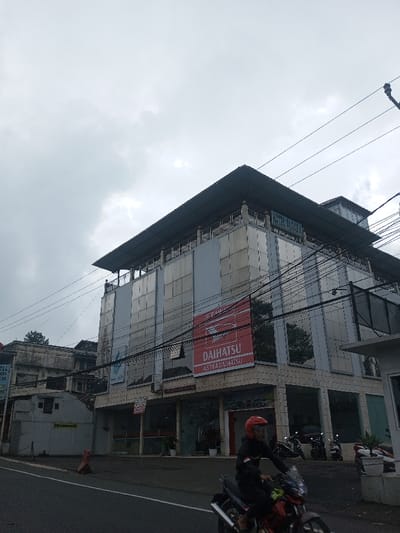A low-angle daytime shot shows a commercial building with a "DAIHATSU ASTRA DAIHATSU" sign. Power lines crisscross the sky. A motorcyclist in a red helmet rides on the street in front of the building, which also features "HOTEL AGRA" signage. Other parked motorcycles are visible. The sky is overcast, suggesting a cloudy or rainy day. A smaller building is partially visible to the left.