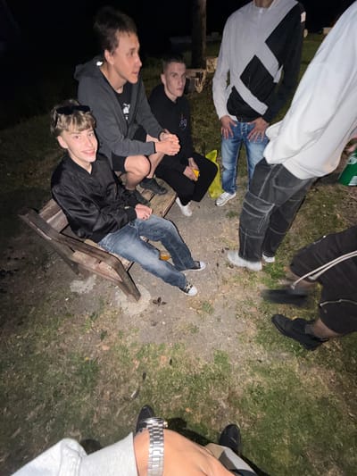 The image shows a group of people in an outdoor setting, likely at night due to the low light conditions. The surface is a mix of dirt, gravel, and grass. At least seven people are visible, most of whom appear to be young adults. Some are sitting on what appears to be a wooden bench or structure. Several are wearing casual clothing, including hoodies, jeans, and sneakers. A wristwatch is visible on the wrist of an individual, and in the background, some buildings or structures can be vaguely discerned. The location is unknown and not locatable with the provided image, and no information leads to finding it.