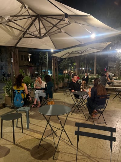 The image depicts an outdoor café scene at night at Kiosko, Hillel Street 18, West Jerusalem, Israel. The paved patio is illuminated by large white square parasols with integrated exposed bulbs, complemented by distant string lights and streetlights. Numerous dark metal bistro tables with round tops and matching folding chairs are arranged across the space.

In the mid-ground left, three individuals are seated at a table: a woman with long dark hair wearing a yellow top and a light blue backpack (back mostly to camera), a woman with short dark curly hair in a light-colored long-sleeved top drinking from a bottle, and a woman with dark curly hair in a dark jacket with an orange-brown shoulder bag hanging from her chair. To their right, a man with short dark hair in a black t-shirt is seated at a separate table, smiling and looking left; a glass and a small metal ashtray are present on his table. Further right, a woman with long dark hair, wearing a dark top and jeans, is seated with her back partially to the camera; a glass is on her table. Additional patrons and a woman in a black blazer standing are visible in the background.

The background features tall trees, dense foliage, and multi-story historic stone buildings on the left. A street with faint distant activity is visible beyond the patio area.