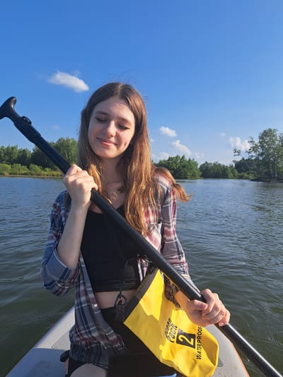 A young woman with long, wavy brown hair and dimples smiles with her eyes closed as she holds a black paddle across her chest. She is wearing a black crop top and a plaid shirt open over it. A yellow waterproof bag is attached to her. She is sitting on a stand-up paddleboard on a body of water. The background shows trees and a bright blue sky with scattered white clouds. This scene is taking place in the city of Razdolnoye, Russia, on a sunny day.