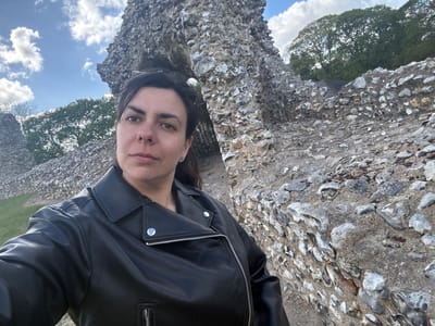 A medium close-up outdoor selfie features a woman looking directly towards the camera with a neutral, slightly serious expression. She has dark hair and dark eyes. She is wearing a black leather jacket with silver zippers and silver snap buttons on the lapels and pockets.
The background primarily consists of an old, weathered stone wall or ruins, likely constructed from flint and mortar, which occupies the left side of the frame and extends into the foreground. The irregular-shaped stones are a mix of light grey and darker hues. To the right of the woman and behind the stone structure, there is a lush green landscape with several large, mature trees boasting full, verdant canopies. Above the trees, a clear blue sky is visible, dotted with scattered white, fluffy clouds.
The scene is brightly illuminated by natural daylight, indicating good weather conditions. There is no specific activity depicted other than posing for the photograph. The nature of the stone construction and the presence of historical-looking ruins in a green setting are consistent with the location context of King's Lynn and West Norfolk, United Kingdom, suggesting a historical site or park. No visible text is present in the image.