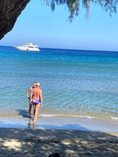 This image captures a bright, daytime coastal scene at a beach in Φάρος, Greece. Two adult males are observed standing in the shallow, clear turquoise waters at the shoreline. The man in the foreground, with light hair, is viewed from behind, wearing blue swim briefs with a prominent white and red design on the rear. He faces away from the viewer and towards a second man, who is partially visible, facing the first, with light hair and wearing lighter-colored swim briefs. Gentle waves break at their feet. In the mid-ground, a large white motor yacht is anchored on the deeper blue sea under a clear blue sky. Several small yellow buoys are visible on the water's surface. The immediate foreground features light-colored sand marked by prominent shadows cast by overhead foliage, which is partially visible on the left and top edges of the frame.