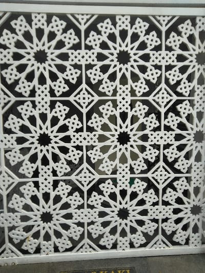 This image captures a close-up of a white, perforated decorative screen or grille, exhibiting an intricate repeating geometric pattern, located in the city of San Cristóbal, Costa Rica. The pattern is composed of interwoven eight-pointed stars and smaller quatrefoil-like motifs, rendered in a style reminiscent of Moorish or Islamic geometric art. The white framework sharply contrasts with the dark, undifferentiated background visible through the cutouts, suggesting an interior or shaded architectural element. The screen is bordered by a plain white frame at the top and sides, and rests on a dark grey horizontal base. Partial, faint text, including the letters "AS KAKI," is visible on this base. No individuals, activities, or interactions are depicted in the scene. The environment beyond the screen is obscured by darkness and the screen's design.