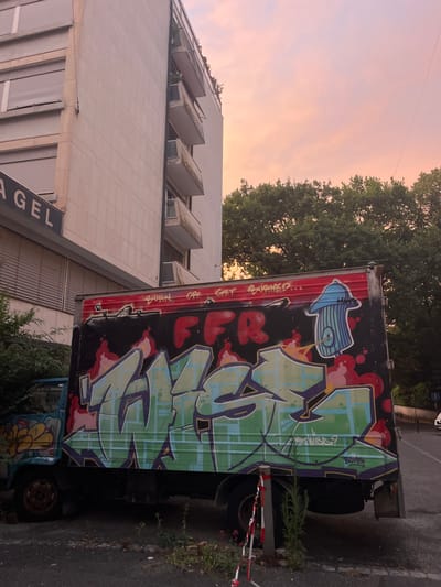 The image depicts a graffiti-covered truck parked next to a modern building. The truck's trailer is adorned with large, colorful graffiti in green, purple, and red. A blue cartoonish figure resembling a hydrant is also visible on the side of the trailer. The building appears to be an office or residential building with a facade of white tiles and a series of angled windows. The sky in the background is a soft pink and orange, suggesting either sunrise or sunset. Trees with green foliage are visible behind the truck and next to the building. There is some red and white striped tape on a pole near the truck, possibly indicating a hazard or restricted area. The overall scene appears to be an urban or suburban setting.

Visible text includes "GET BURNED..." in red spray paint on the side of the truck, and smaller text within the graffiti, some of which is illegible. There is also text on a sign on the building that reads "AGEL", part of what is likely a larger word. The weather appears clear. The scene conveys an atmosphere of urban decay or artistic expression within an otherwise unremarkable environment. The orientation of the image is such that the ground appears on the right side of the frame and the sky on the left.