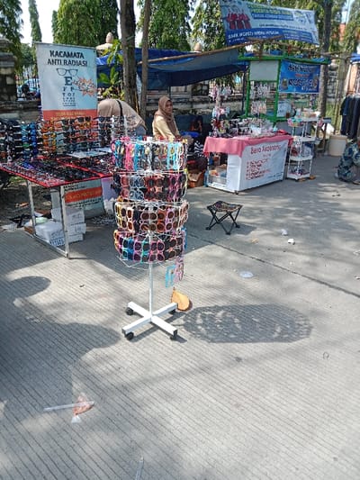 The image shows a street stall selling sunglasses in Kendal, Indonesia. The stall is located near a large tree and a blue tent. There are several pairs of sunglasses displayed on a rotating stand in the center of the image. The stand is white and has a cross-shaped base. There are four wheels on the base and the sunglasses are arranged in a circular pattern on the stand. There are various colorful sunglasses on the rotating display. To the left, there are two tables displaying sunglasses, one with a sign that says "Kacamata Anti Radiasi" and the other with a sign that says "Rames Protection". There is a woman wearing a hijab and a white shirt standing behind the sunglasses table on the left. The stall is surrounded by a concrete area and there is a large tree in the background. There is a small stool on the right side of the image. The environment is an urban area with a sidewalk and a street. There are some pieces of trash on the ground. The ground is grey and made of concrete. There is also a white plastic bag and a brown leaf on the ground. The sky is partly cloudy and the weather is sunny.