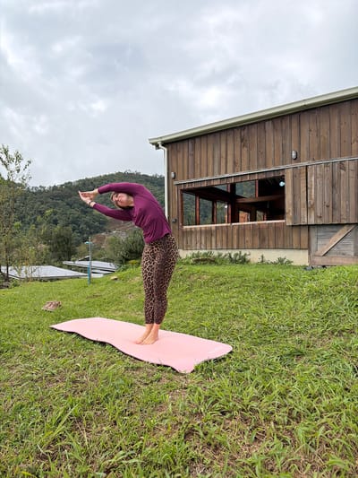 A woman with blonde hair is performing a yoga pose outdoors in Ban Khun Wang, Thailand, on a cloudy day. She is wearing a long-sleeved purple shirt and leopard-print leggings. She stands on a pink yoga mat placed on a grassy hill. Her arms are raised above her head, interlaced, and her body is arched backward. Behind her is a wooden building with a large window, and in the distance, there are lush green hills covered in trees. A pair of tan sneakers are visible on the grass to the left of the yoga mat.